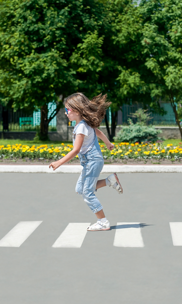 Ein Mädchen läuft über einen Zebrastreifen. Sie trägt einen hellblauen Overall und weiße Sandalen, ihr langes Haar weht im Lauf. Im Hintergrund sind grüne Bäume und ein Blumenbeet zu sehen.