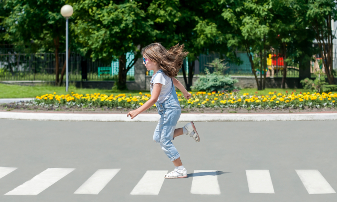 Ein Mädchen läuft über einen Zebrastreifen. Sie trägt einen hellblauen Overall und weiße Sandalen, ihr langes Haar weht im Lauf. Im Hintergrund sind grüne Bäume und ein Blumenbeet zu sehen.