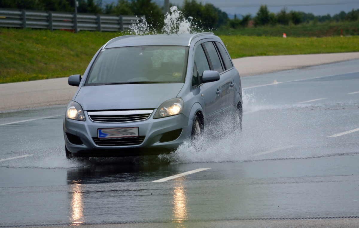 Ein silbernes Auto fährt bei Regen über eine nasse Fahrbahn, wodurch Wasserfontänen von den Reifen aufgespritzt werden.