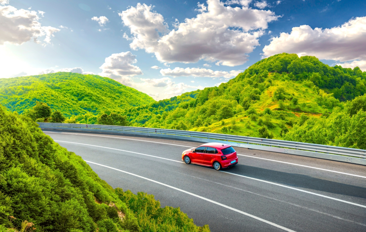 Ein rotes Auto fährt auf einer kurvigen Landstraße durch eine grüne Hügellandschaft unter blauem Himmel mit weißen Wolken.