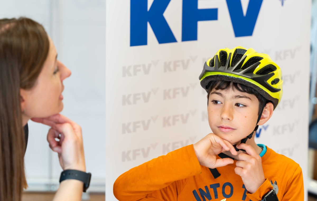 Ein Junge mit orangem Pullover und gelbem Fahrradhelm schließt den Kinnriemen seines Helms. Im Hintergrund ist ein Banner mit dem Schriftzug KFV zu sehen.