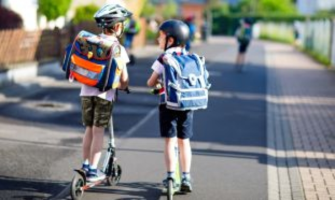 Zwei Schulkinder mit Rucksäcken und Helmen fahren mit Tretrollern nebeneinander auf dem Gehweg zur Schule.