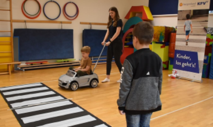 In einer Turnhalle üben Kinder spielerisch Verkehrssicherheit: Ein Kind sitzt in einem kleinen Spielauto, ein anderes beobachtet, und eine Erwachsene zieht das Auto über einen auf dem Boden ausgelegten Zebrastreifen.
