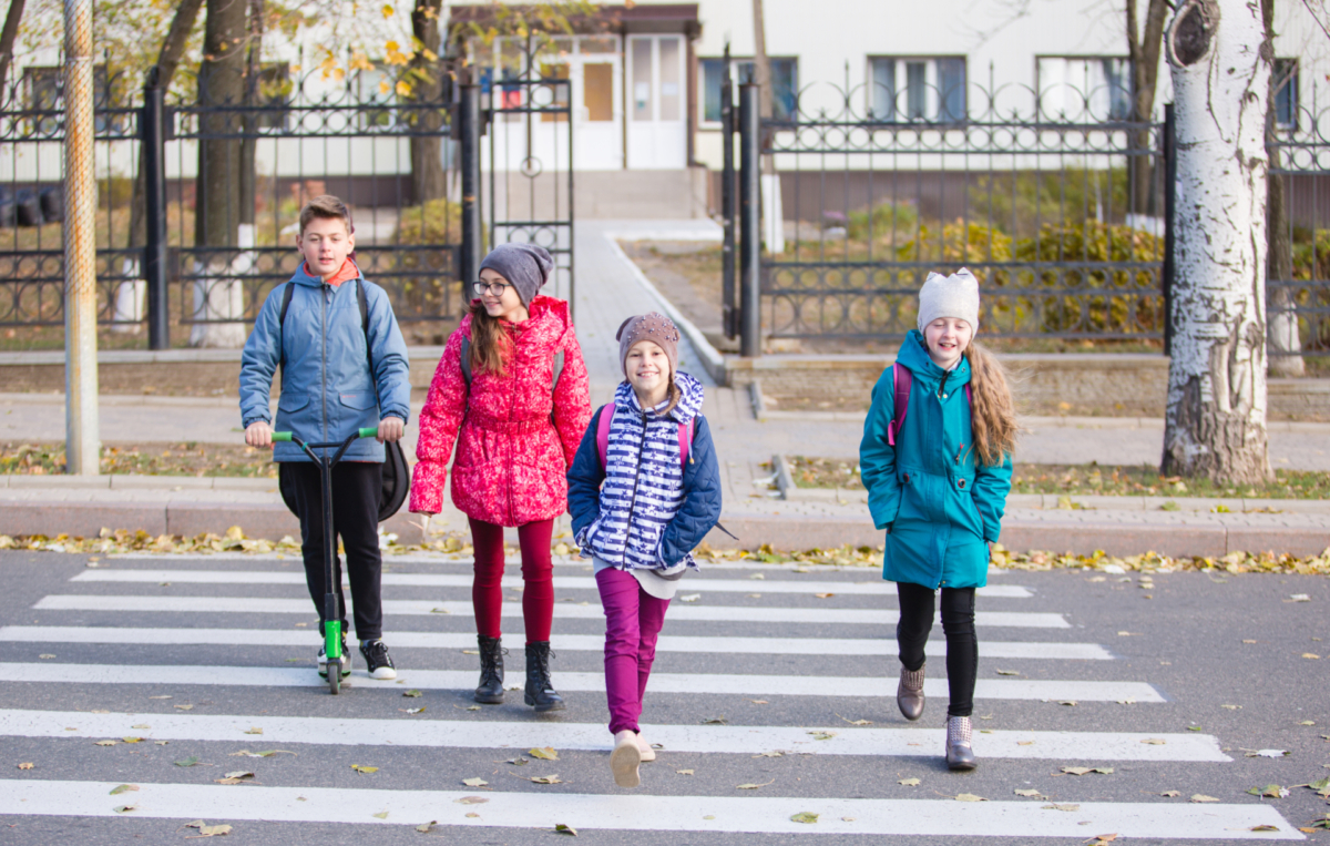 Vier Kinder mit Rucksäcken überqueren gemeinsam einen Zebrastreifen. Eines fährt mit einem Roller, die anderen drei gehen nebeneinander. Alle tragen bunte Jacken und Mützen, im Hintergrund ist ein Gebäude mit Zaun zu sehen.