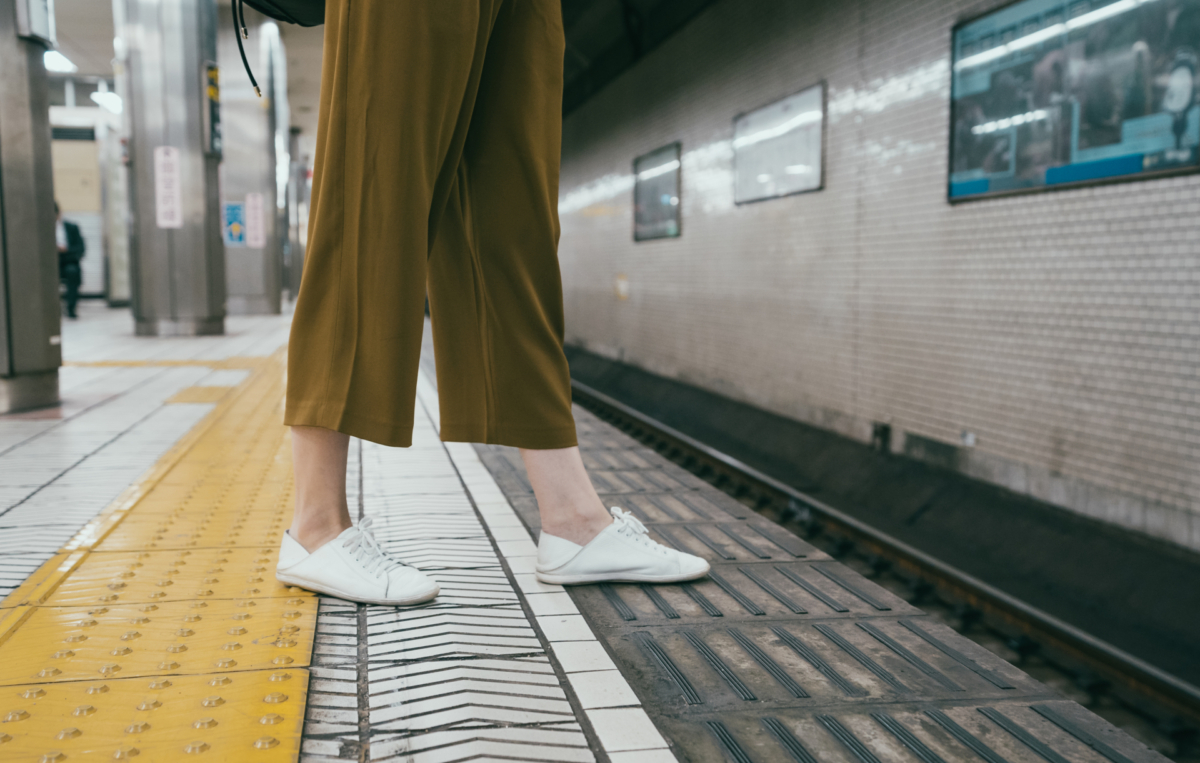 Person mit weißen Sneakers und brauner Hose steht dicht am Bahnsteigrand einer U-Bahn-Station, nahe den Sicherheitsmarkierungen.
