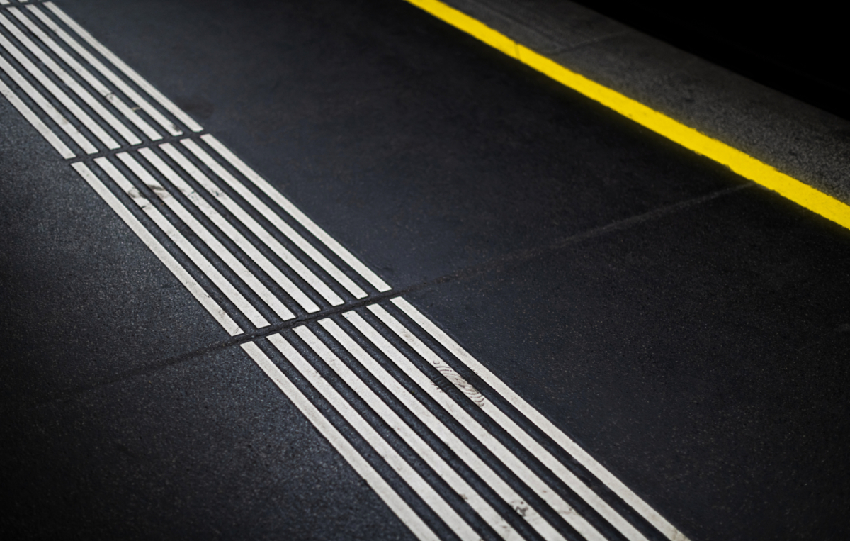Bodenmarkierungen auf einem Bahnsteig: weiße taktile Leitstreifen für sehbehinderte Menschen verlaufen parallel, daneben ist eine gelbe Sicherheitslinie am Bahnsteigrand zu sehen.