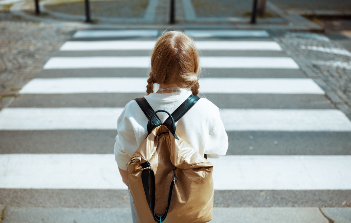 Ein Kind mit Rucksack steht vor einem Zebrastreifen und wartet, um die Straße zu überqueren.