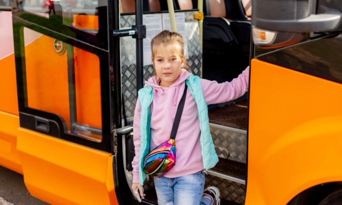 Ein Kind steigt in einen orangefarbenen Bus ein und hält sich am Türrahmen fest, während es in die Kamera blickt.