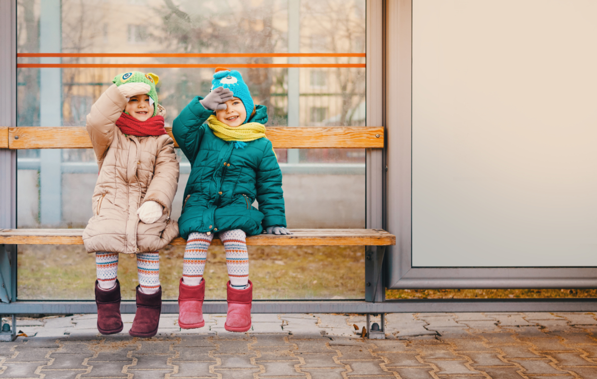 Zwei Kinder in Winterkleidung sitzen lachend auf einer Holzbank an einer Bushaltestelle, beide tragen bunte Mützen, Schals und Stiefel.