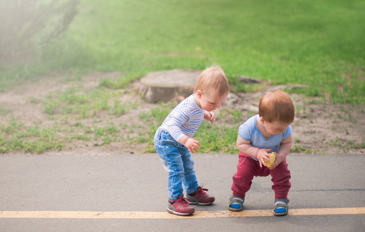 Zwei Kleinkinder spielen gemeinsam auf einem Gehweg; eines beugt sich nach vorne, während das andere ein Spielzeug in den Händen hält.