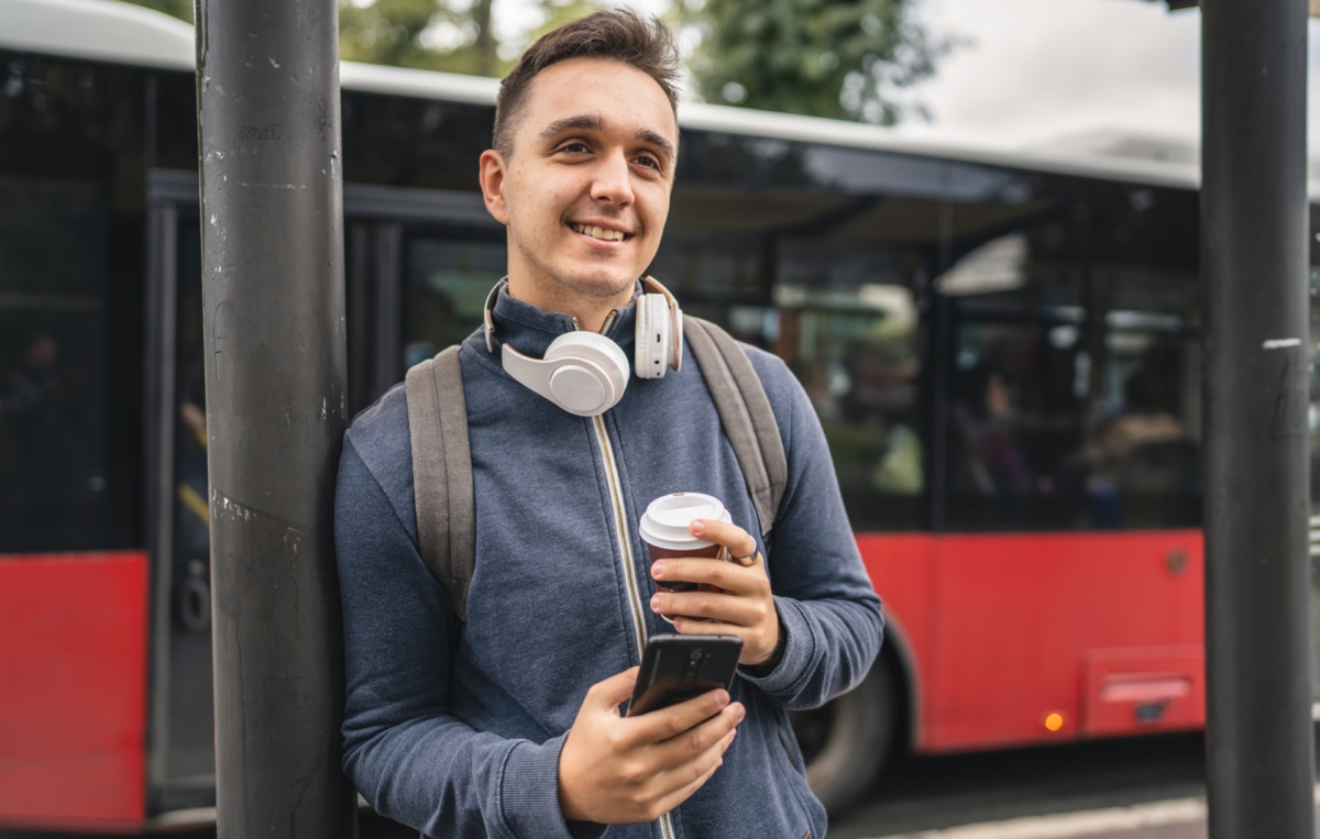 Ein junger Mann mit Rucksack und Kopfhörern um den Hals steht an einer Bushaltestelle, hält einen Kaffeebecher in der einen und ein Smartphone in der anderen Hand, während im Hintergrund ein Bus zu sehen ist.
