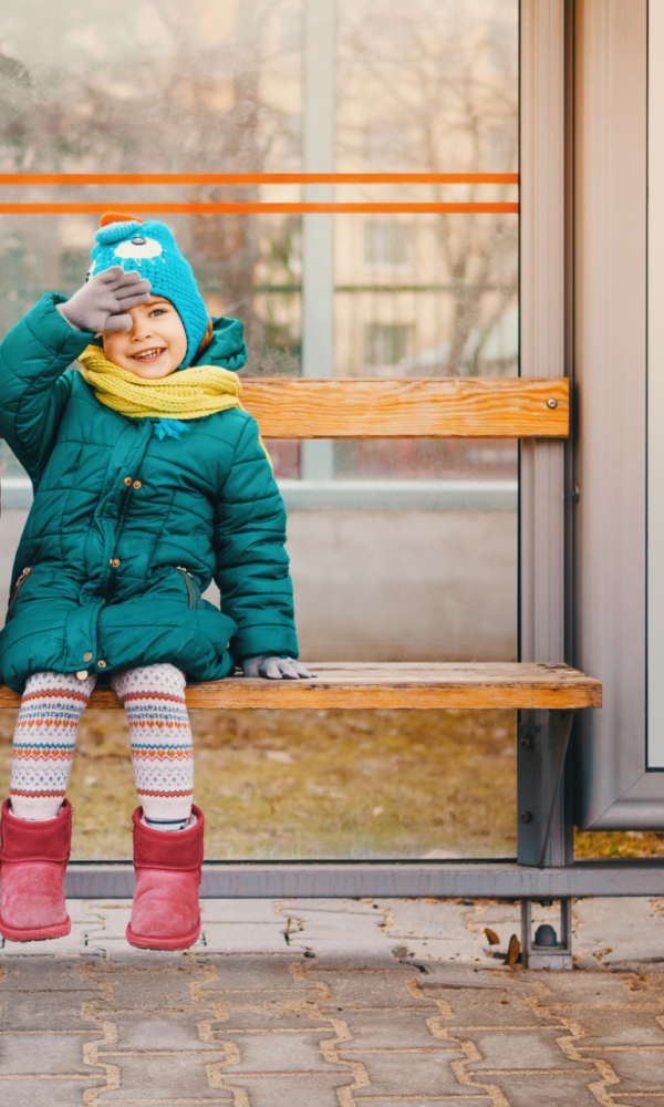Zwei Kinder in Winterkleidung sitzen lachend auf einer Holzbank an einer Bushaltestelle, beide tragen bunte Mützen, Schals und Stiefel.