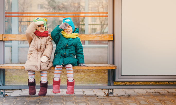 Zwei Kinder in Winterkleidung sitzen lachend auf einer Holzbank an einer Bushaltestelle, beide tragen bunte Mützen, Schals und Stiefel.