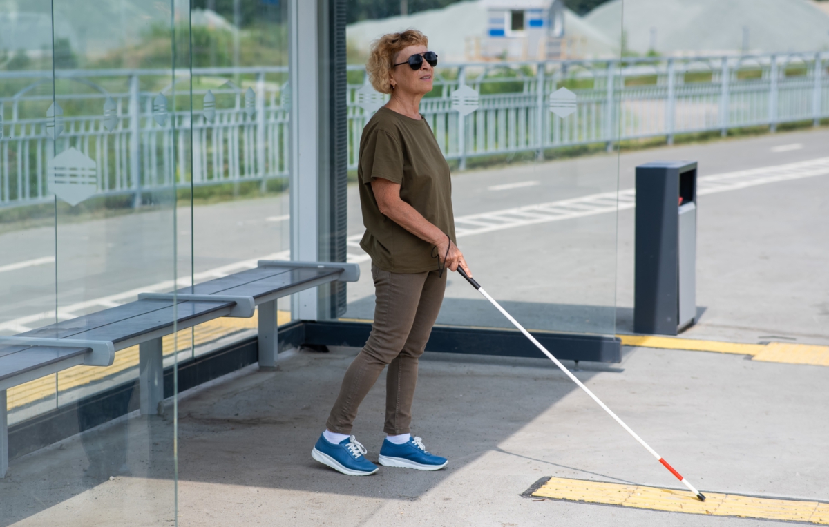 Eine sehbehinderte Frau mit Sonnenbrille steht an einer Haltestelle und benutzt einen weißen Blindenstock zur Orientierung.