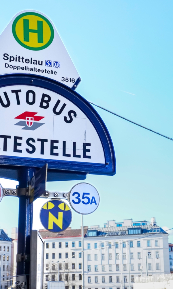 Bushaltestellenschild „Autobus Haltestelle“ in Wien bei der Station Spittelau, mit Linienangaben und Gebäuden im Hintergrund unter blauem Himmel.