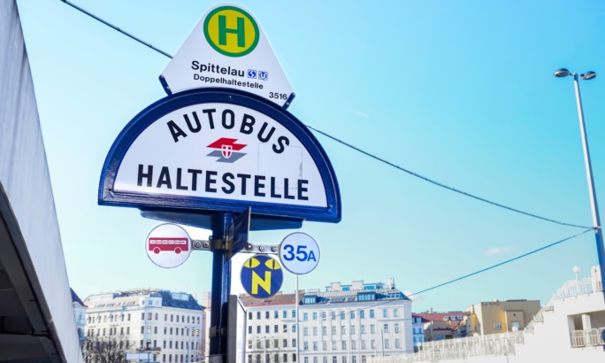 Bushaltestellenschild „Autobus Haltestelle“ in Wien bei der Station Spittelau, mit Linienangaben und Gebäuden im Hintergrund unter blauem Himmel.