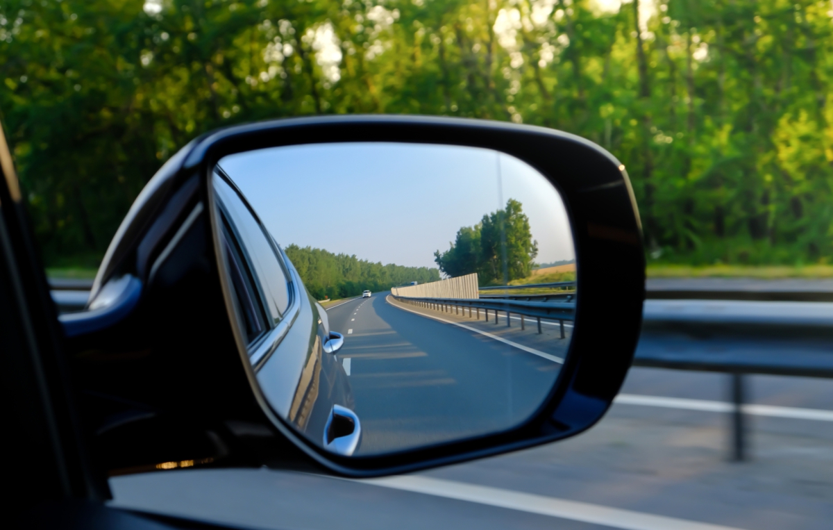 Blick in den Außenspiegel eines Autos, der eine leere Landstraße mit Leitplanken und Bäumen im Hintergrund zeigt.