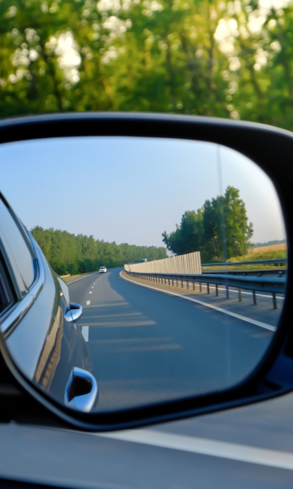 Blick in den Außenspiegel eines Autos, der eine leere Landstraße mit Leitplanken und Bäumen im Hintergrund zeigt.