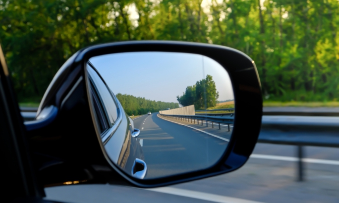 Blick in den Außenspiegel eines Autos, der eine leere Landstraße mit Leitplanken und Bäumen im Hintergrund zeigt.